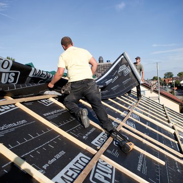 This photograph shows two roofing tradespeople installing SOLITEX Mento Plus membranes on a roof, against a background of a blue sky.